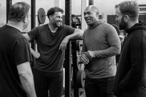 Four men talk and laugh together beside a squat rack